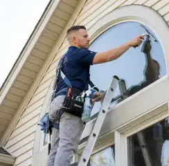 Professional window cleaner on a ladder using a squeegee to clean an exterior arched window, Jackson, MI.